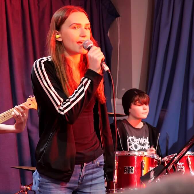 Two teenage girls singing and playing drums at a Junior Academy of Modern Music JAMM session